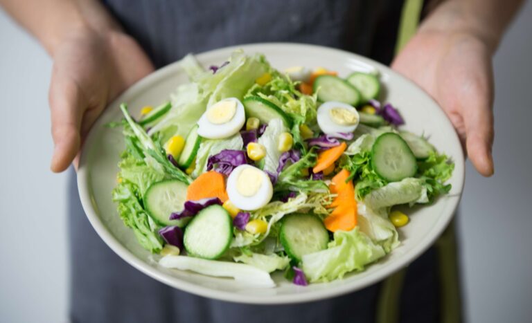 A person holds a plate of fresh salad with greens, cucumbers, boiled egg, corn, and cabbage.