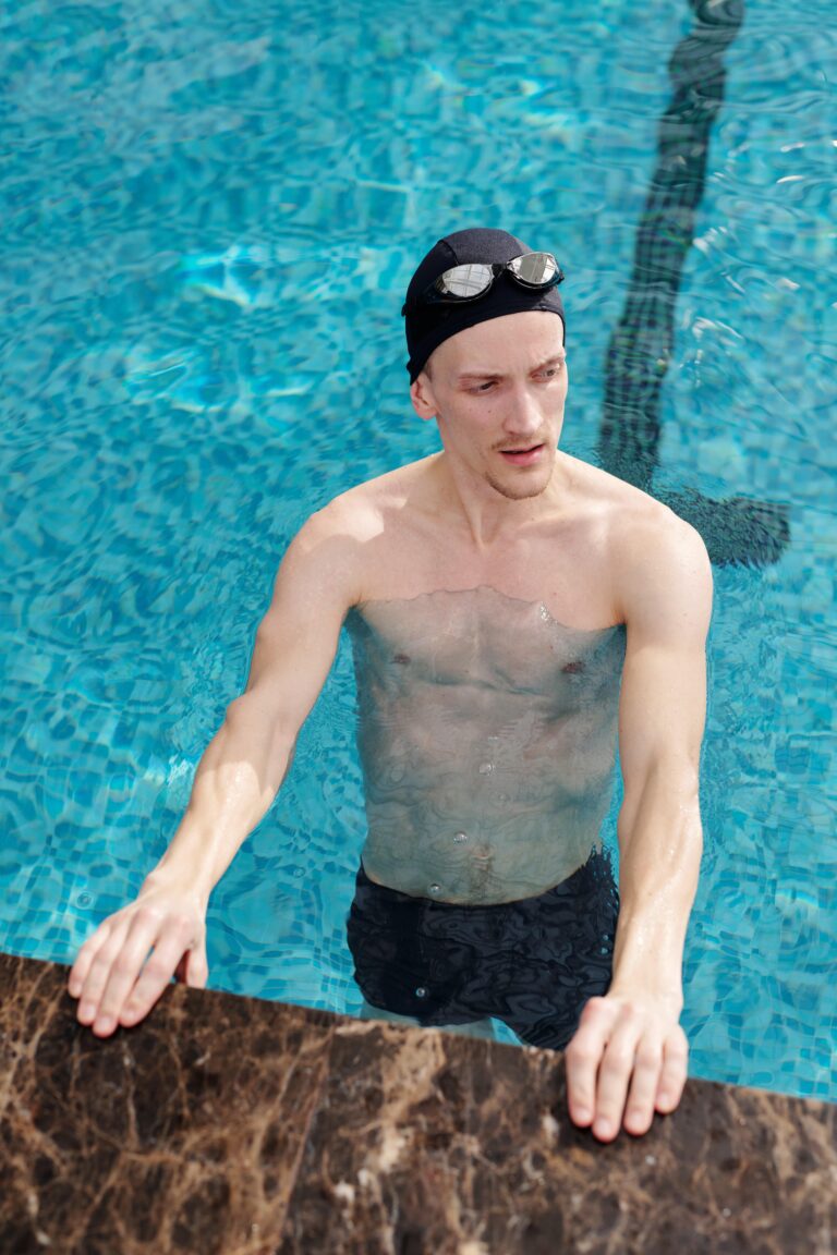 A man in a swim cap and goggles stands in a pool, resting his hands on the edge.