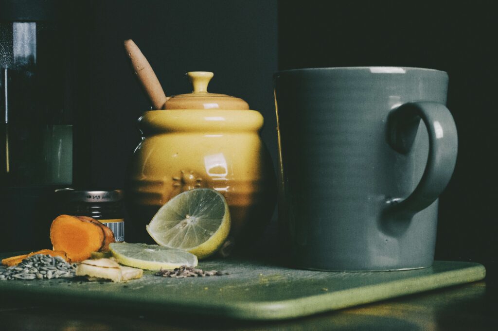 A dark mug on a cutting board with lemon, turmeric, seeds, honey jar, and spice jar.