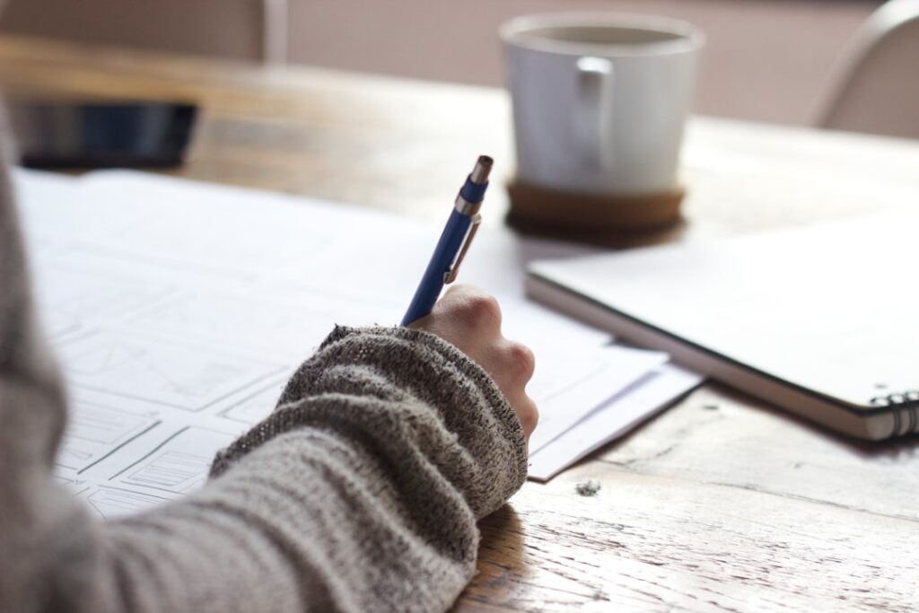 A person in a gray sweater writes on paper at a rustic wooden table. Art of Navigating Healthcare