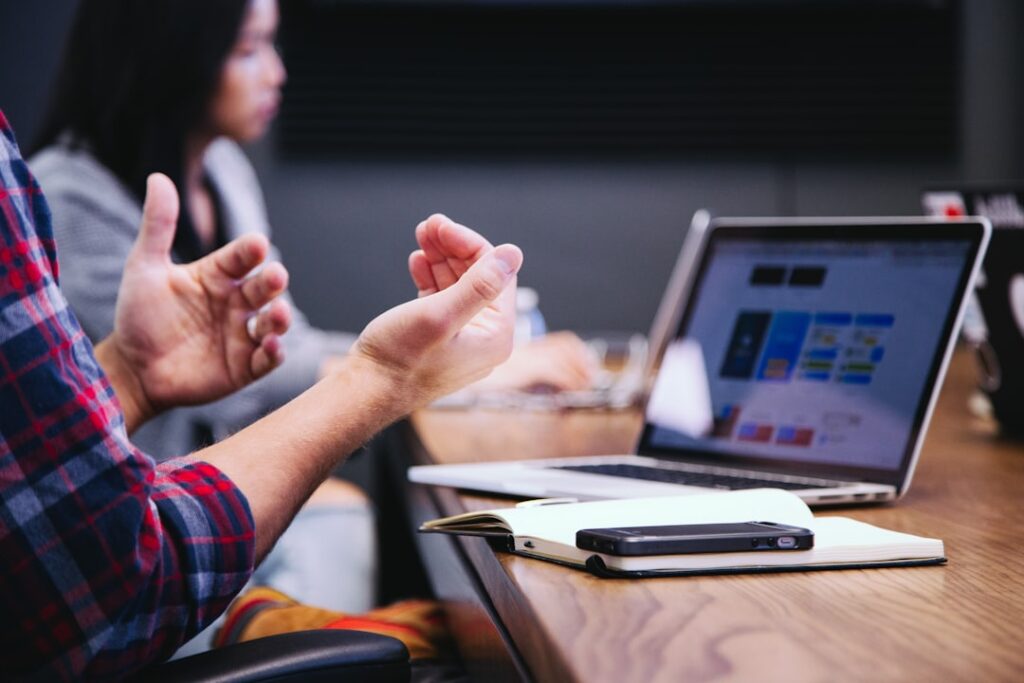 A person gestures at a meeting table with a laptop, notebook, phone. Patient Communication Skills
